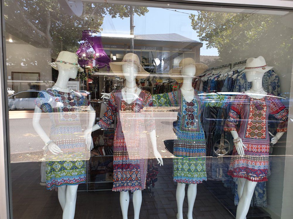 Four mannequins in a shop window display dresses with patterned designs and straw hats.