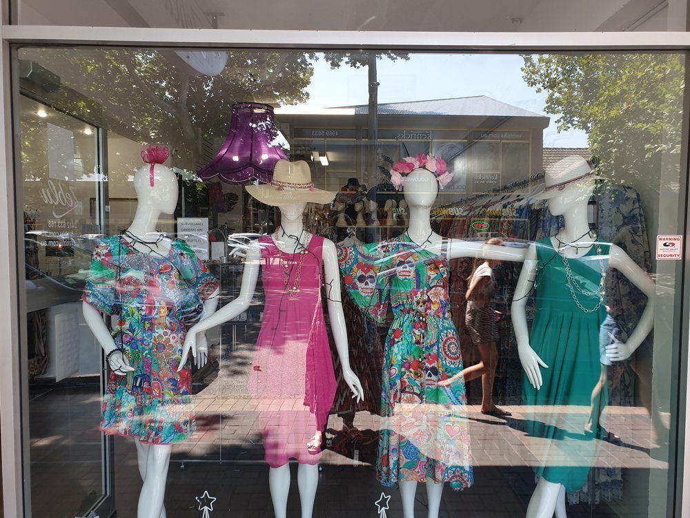 Four mannequins in a store window display colorful dresses with hats and jewelry; reflected street scene visible.