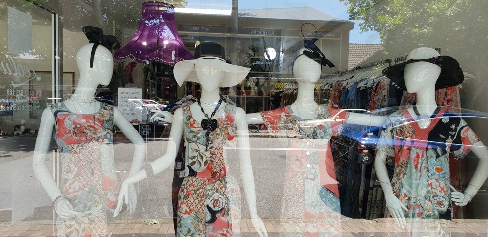 Window display with mannequins wearing floral dresses, hats. Storefront reflection visible.