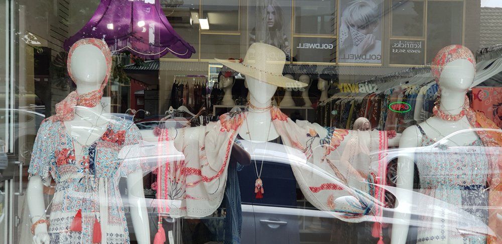 Three mannequins wearing dresses in a store window. The center mannequin is wearing a hat and shawl.