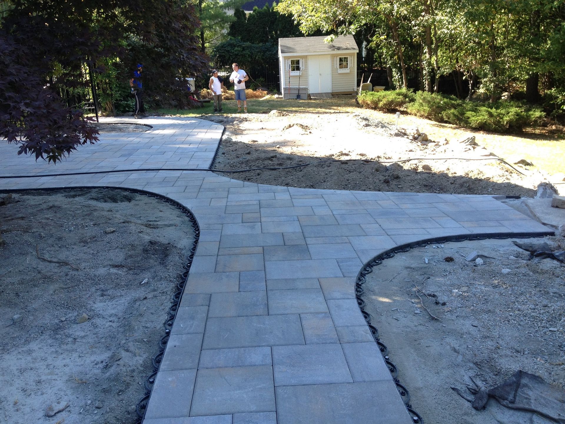 Paver pathway in progress, branching into two directions. People working in the background, small shed.