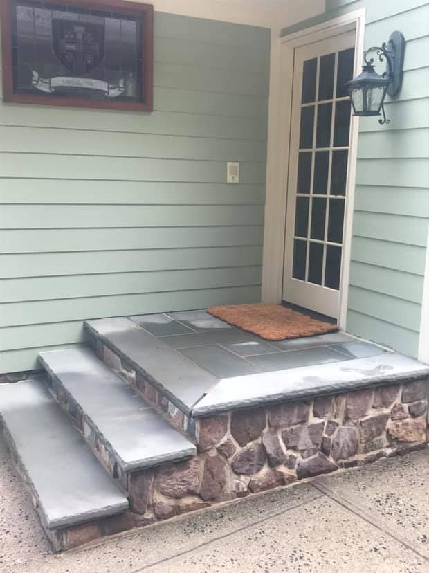 Stone steps leading up to a door with a doormat. Light green siding and a framed crest are visible.