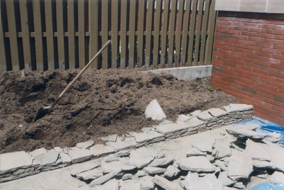 Pile of dirt and shovel in a garden bed with a stone border and a wooden fence.