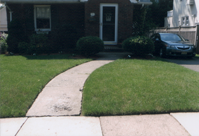 Curved concrete path leading to a house with dark door, flanked by green lawn and a car.