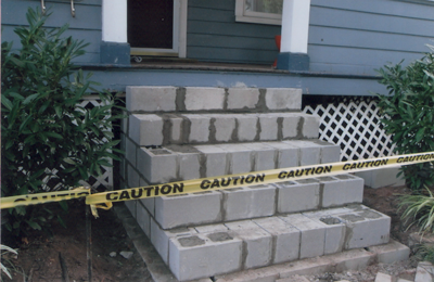 Concrete steps under construction in front of a blue house, with caution tape.