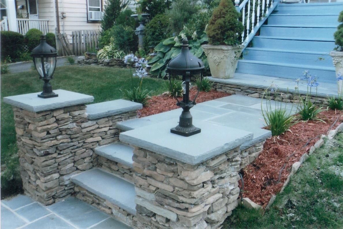 Stone steps and pillars with black lanterns leading to a blue-painted house entrance, set in a landscaped yard.