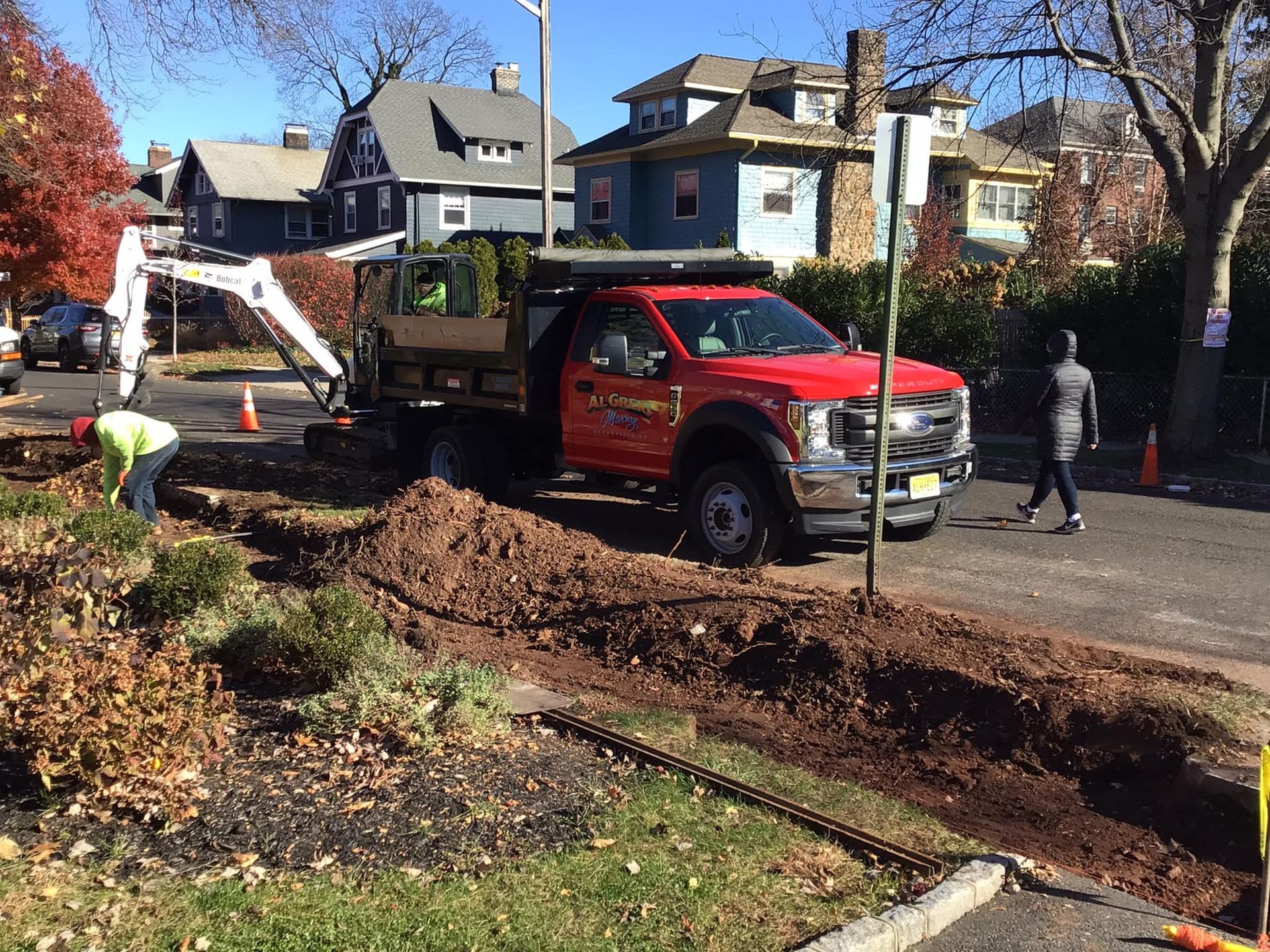 Construction on a street: a red truck, small excavator, workers digging, and a woman walking by.