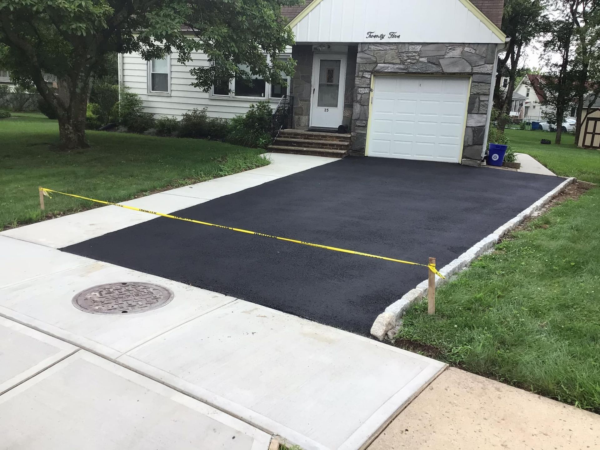 Freshly paved black asphalt driveway bordered by concrete, with yellow caution tape, leading to a house.