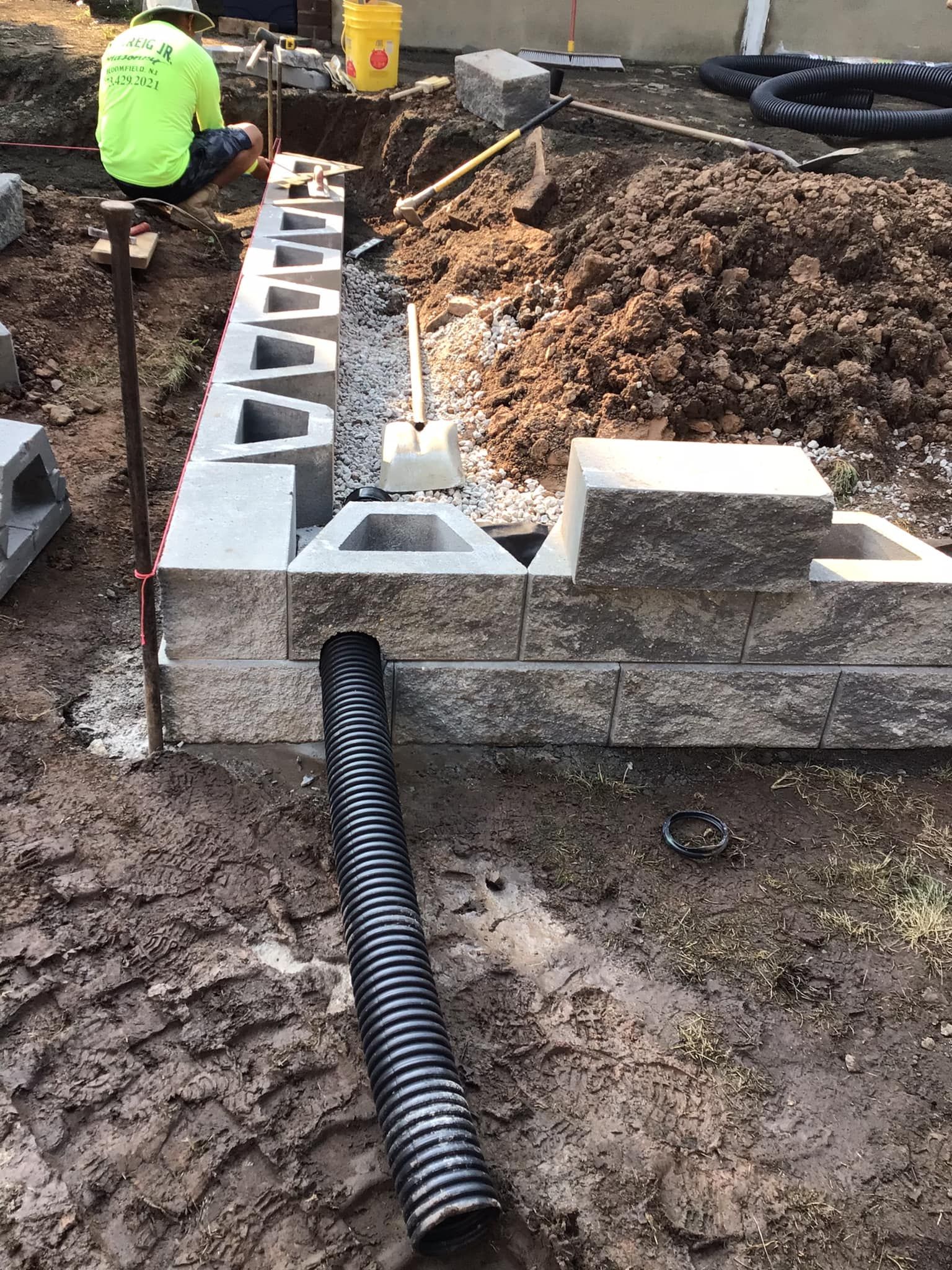 Construction of a retaining wall: worker in safety vest, blocks, drainpipe, dirt, tools, and materials.