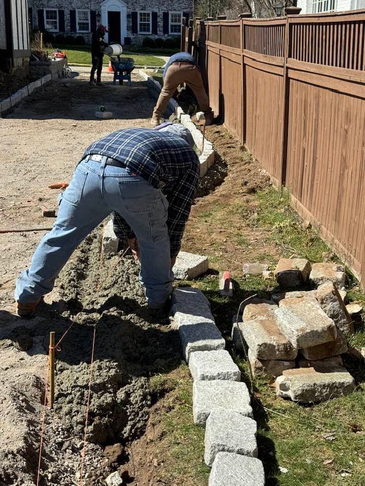 Three workers laying stones along a brown fence and a dirt path; sunny outdoor setting.