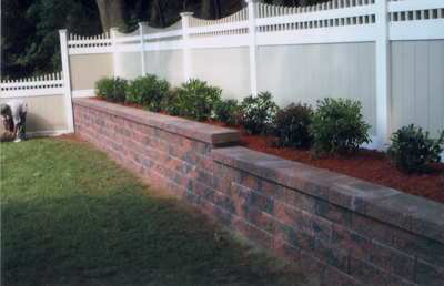 Brick retaining wall with landscaping and white fence.