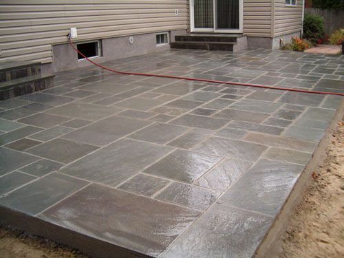 Patio with dark gray stone tiles next to a house with a sliding glass door. A red hose lays on the tiles.