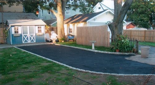 Backyard with asphalt driveway, blue shed, light-colored garage, and trees; person walking near the driveway.
