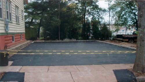 Newly paved asphalt driveway with caution tape in front of a house, trees, and sidewalk.