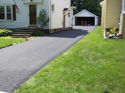 Asphalt driveway leading to a garage, beside a grassy lawn and a house.