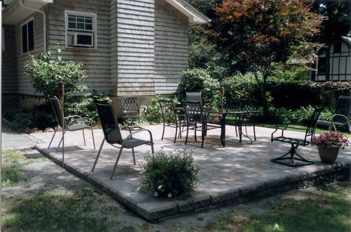 Patio with outdoor furniture next to a gray house, surrounded by grass and greenery.