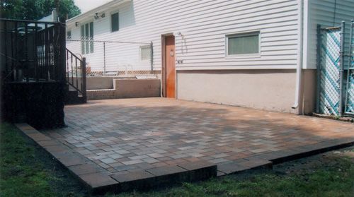 Patio with brick pavers next to a house with wooden deck and a door.