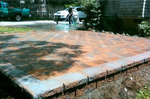 Brick patio being hosed down in front of a house. A person hoses down the surface, a green truck parked on the left.