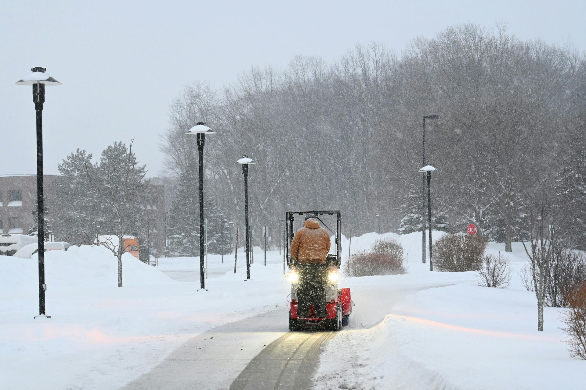Person operating a red snowblower, clearing a snowy path. Snow falling, trees, streetlights.