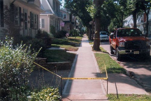 Sidewalk in residential area, yellow caution tape blocking access. Red truck parked on street, trees lining the street.