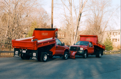 Two red dump trucks parked outdoors, near bare trees and a building.