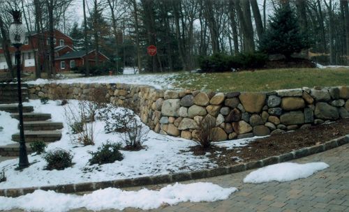 Stone wall bordering a snow-covered lawn and driveway, with a house in the background.