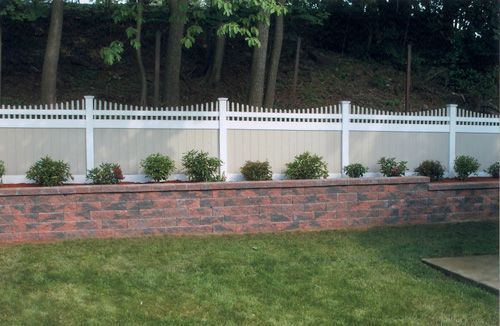 Backyard with brick retaining wall, green lawn, bushes, and white picket fence against a treed hill.