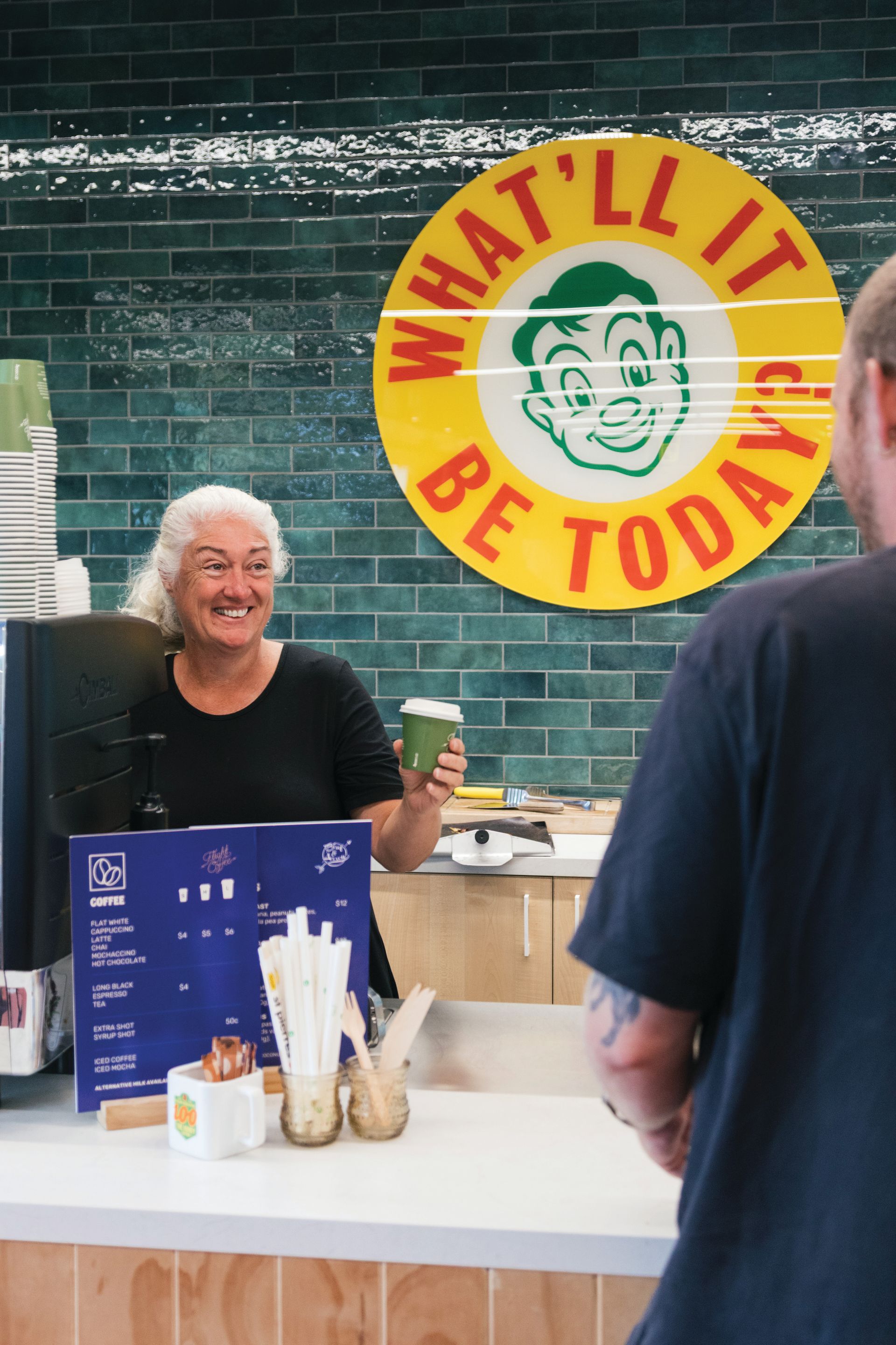 A woman is standing at a counter in front of a sign that says what 'll it be today.