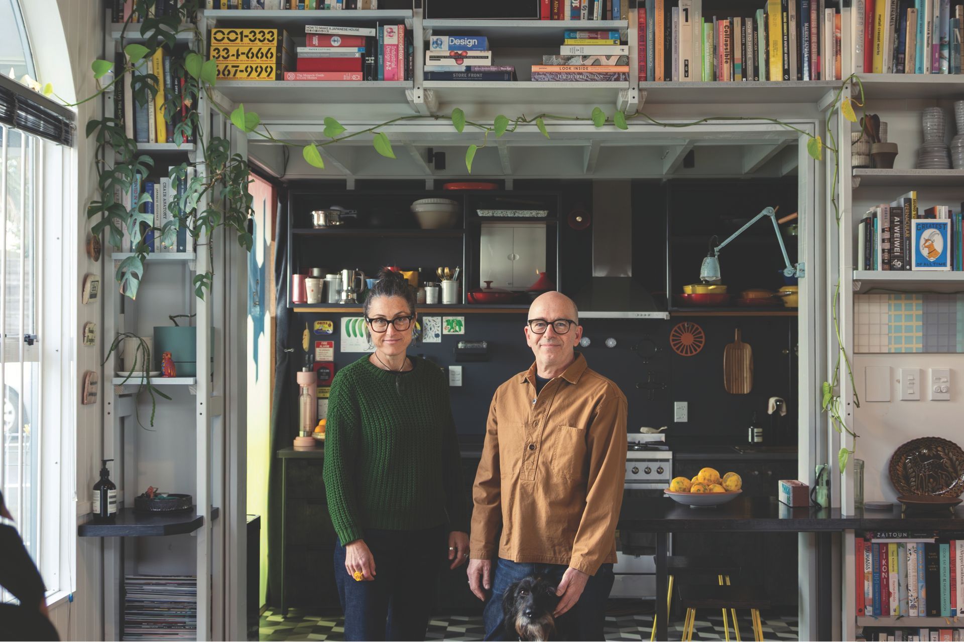 Couple standing in front of a black and white tiled kitchen. Shelves filled with items flank them.