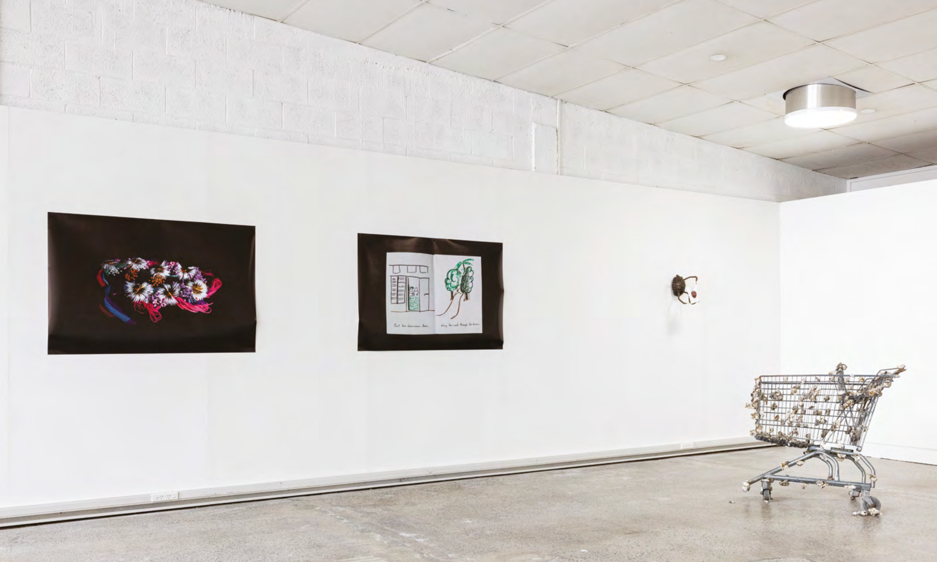A gallery space with art on the walls and a shopping cart in the middle of the room.
