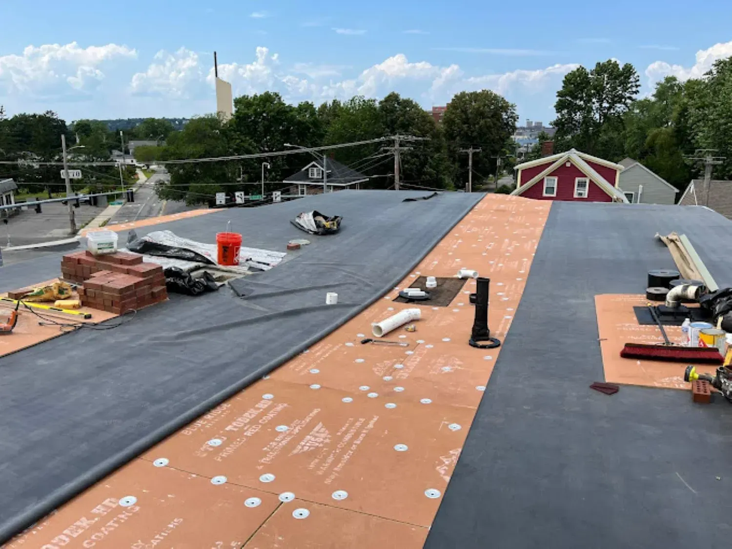 A roof is being installed on a building with a red house in the background.