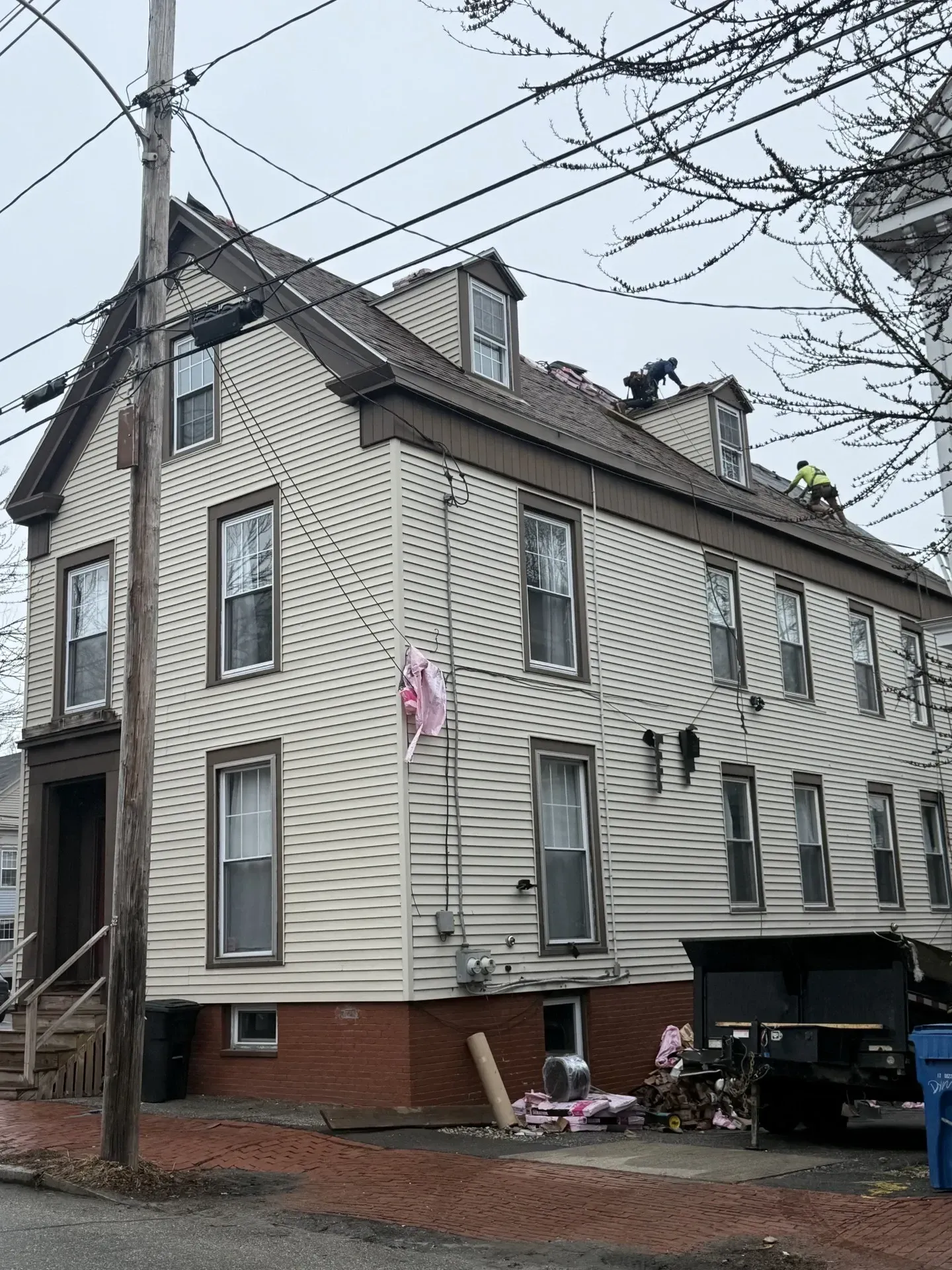 Three-story gray house with brick foundation and dormer windows beside a utility pole and overhead wires.