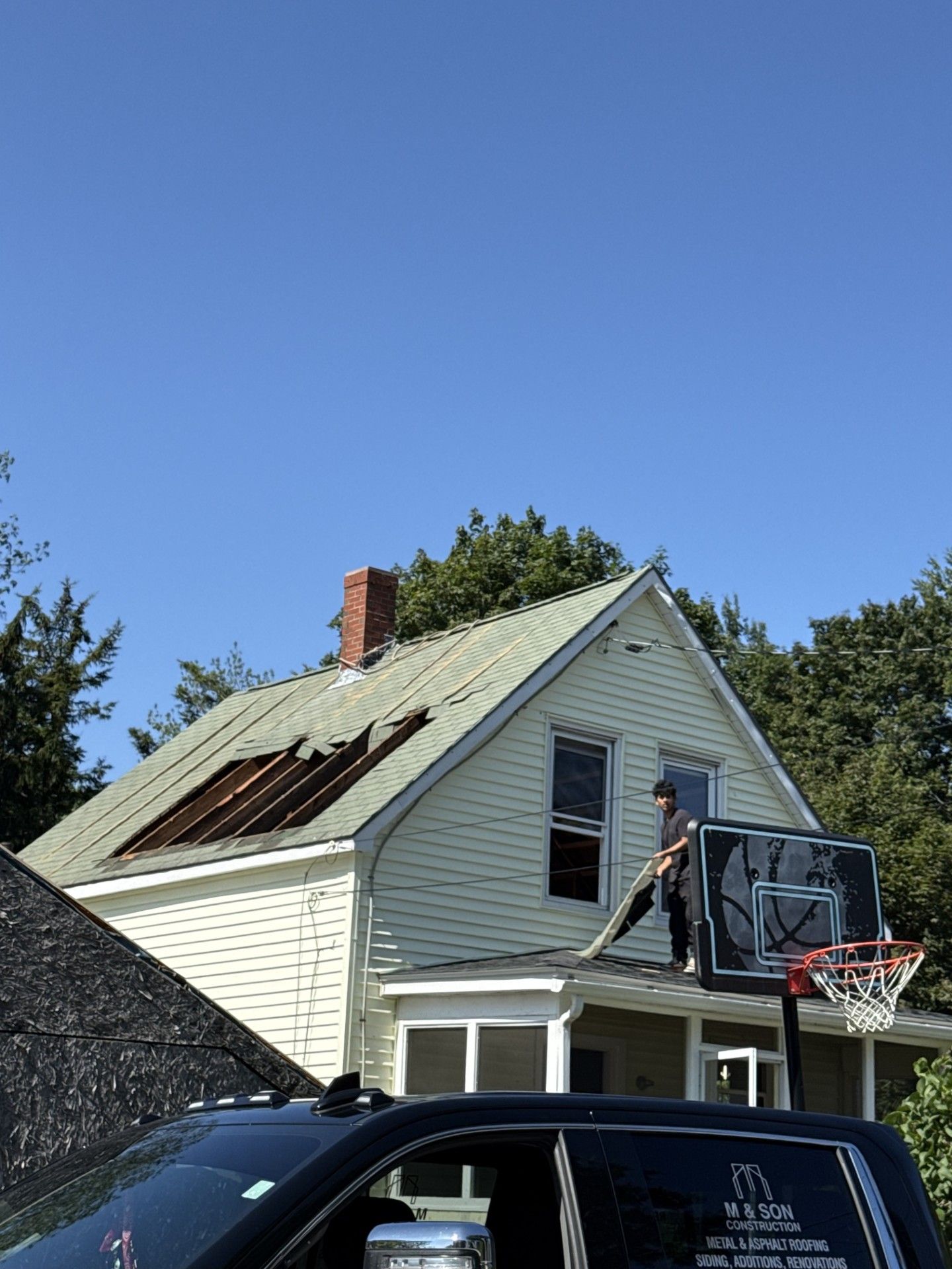 Person on roof of house, removing roof material. Basketball hoop in foreground, blue sky.
