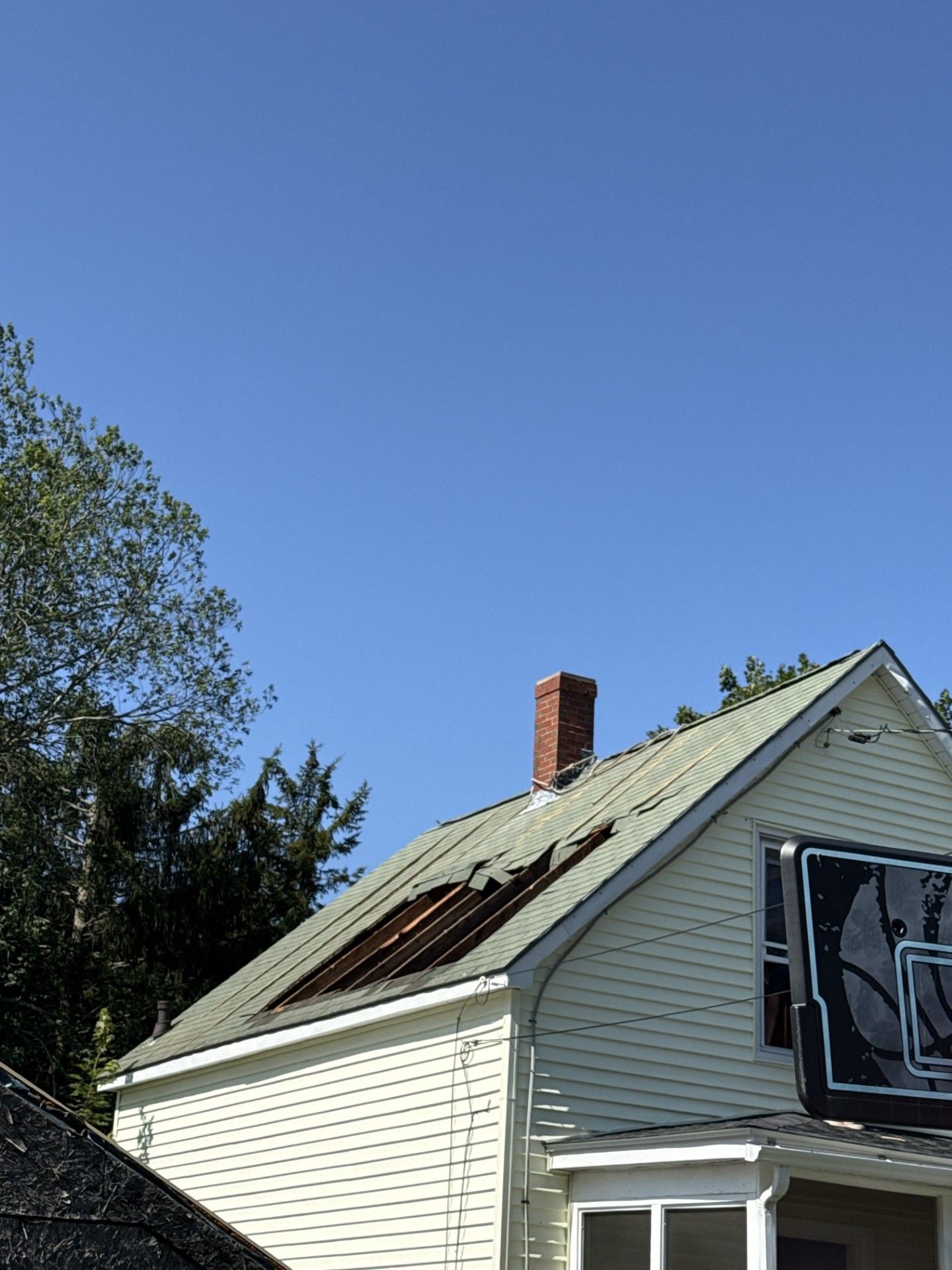 Damaged house roof with missing sections, brick chimney, and clear blue sky.