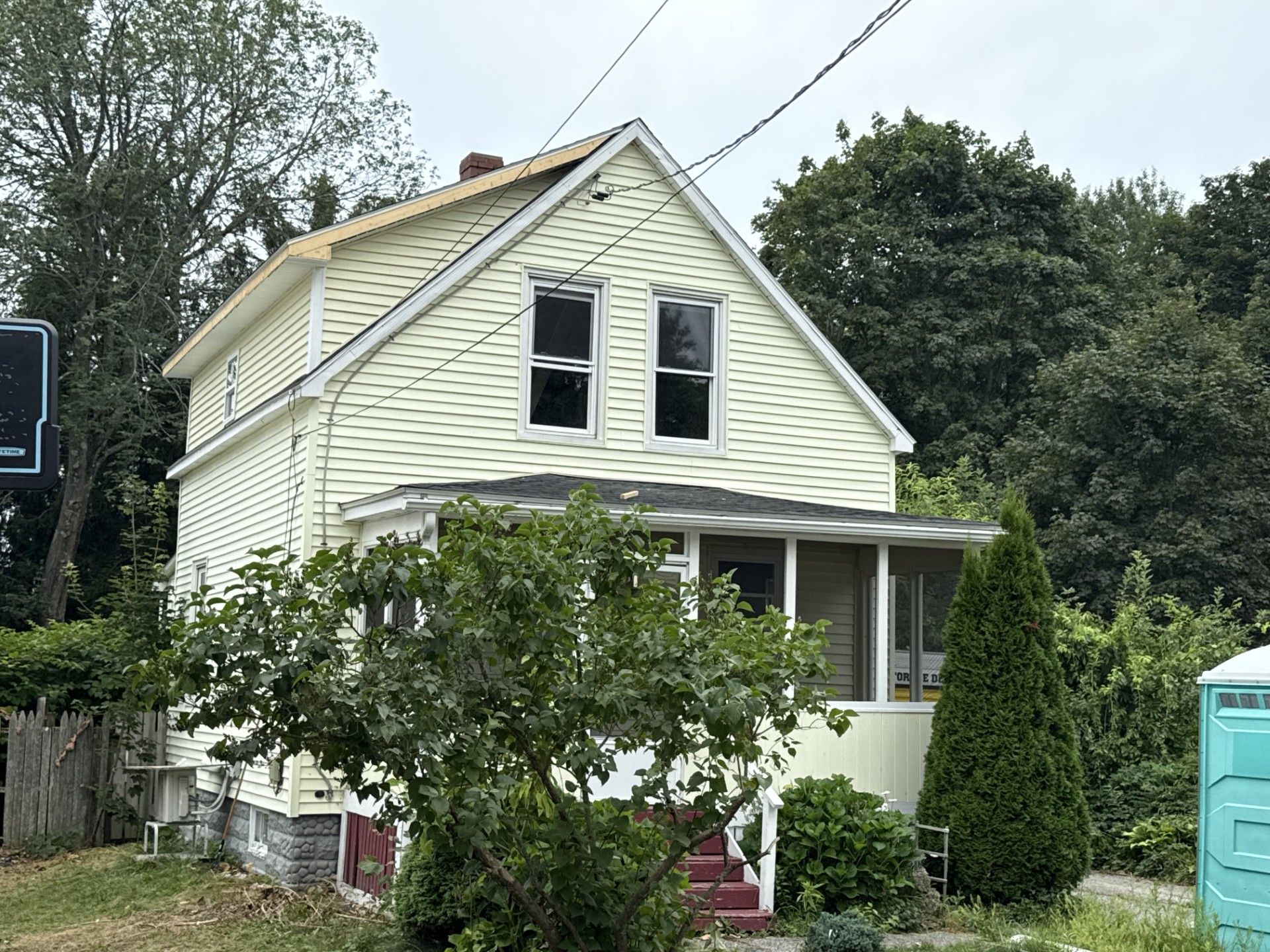 Two-story yellow house with a porch and overgrown yard, cloudy day.
