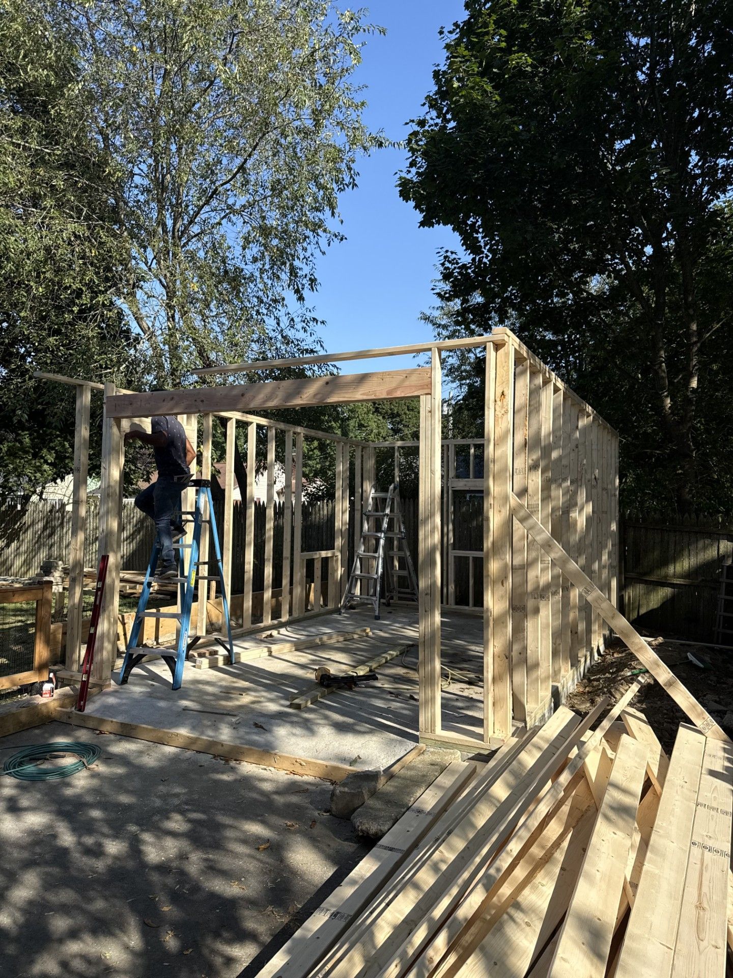 Wooden shed under construction; person on ladder, framing walls, sunlight.