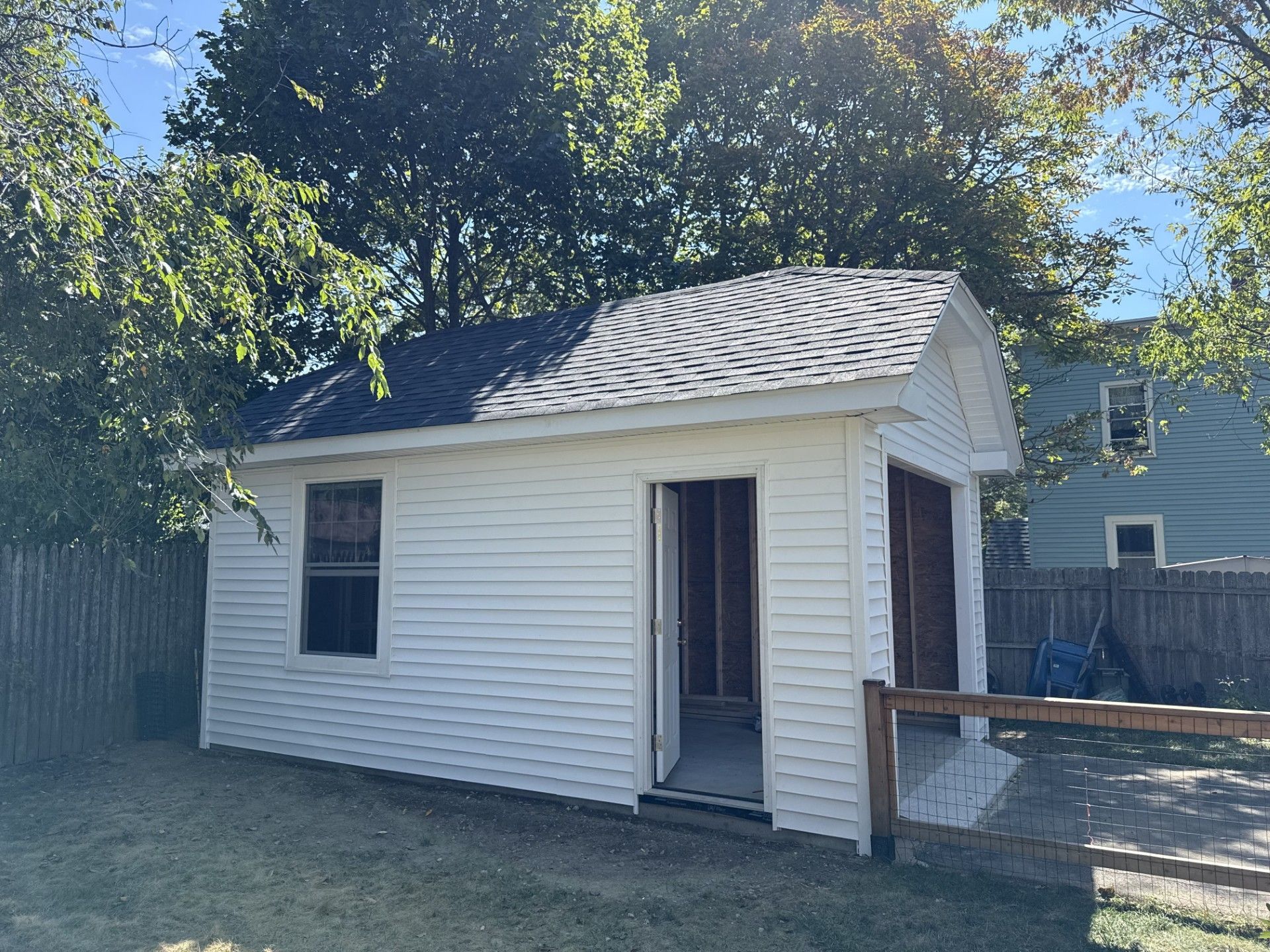 White shed with open door and window, surrounded by green grass and trees, against a blue sky.