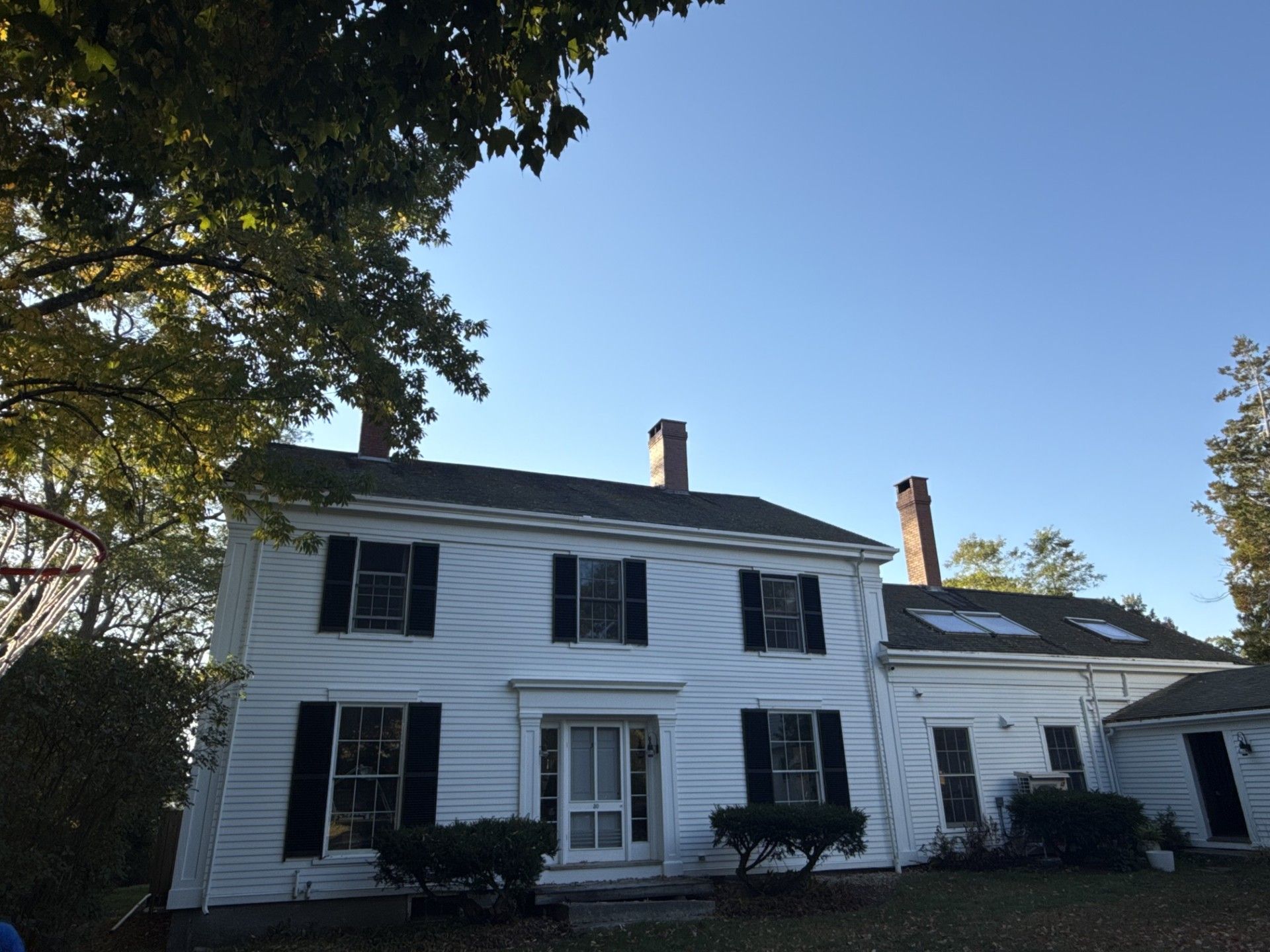 White two-story house with black shutters, multiple chimneys, under a blue sky, surrounded by trees.