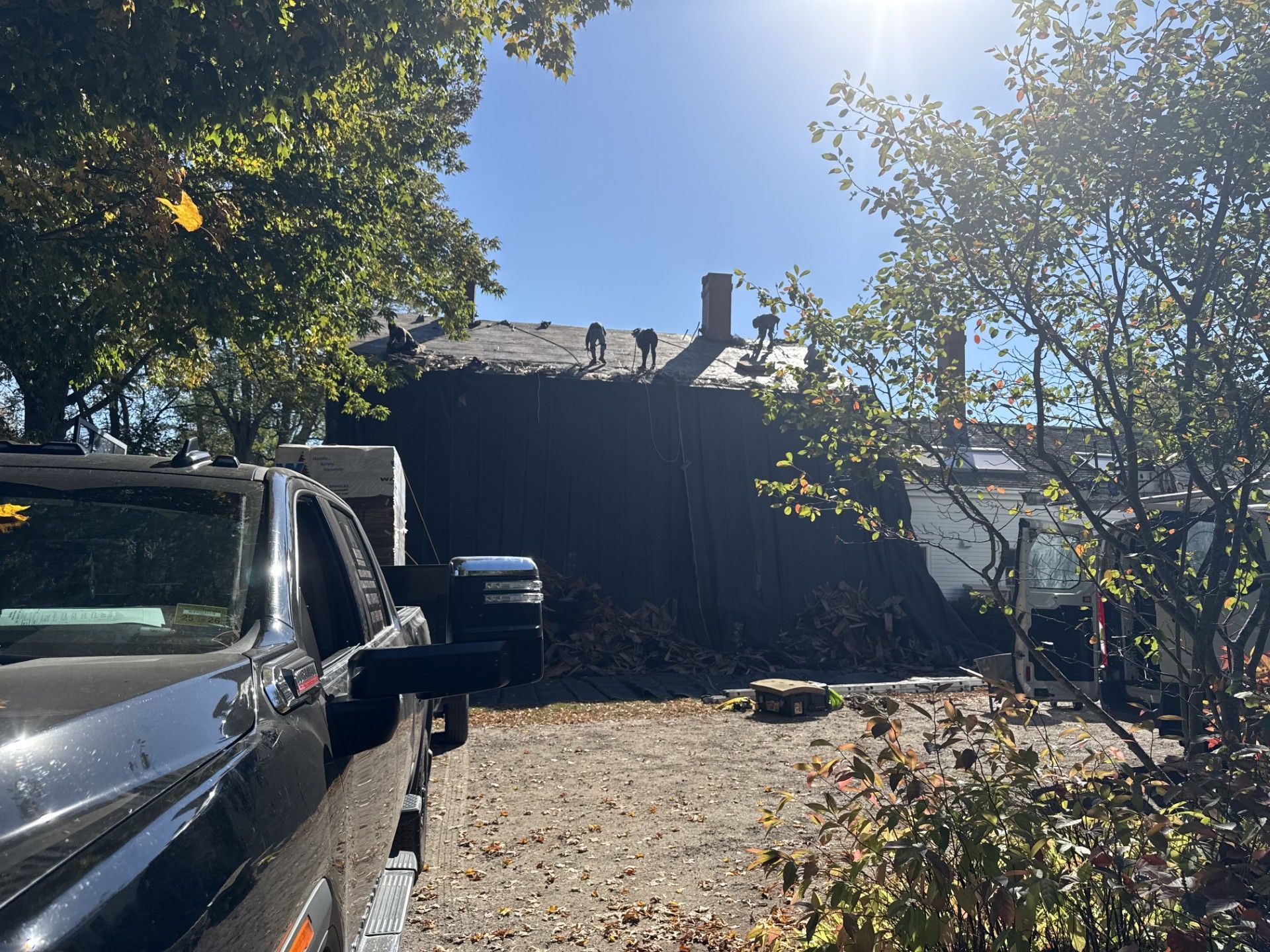 Workers on a rooftop, possibly doing repairs. Black truck parked in front; trees and blue sky background.