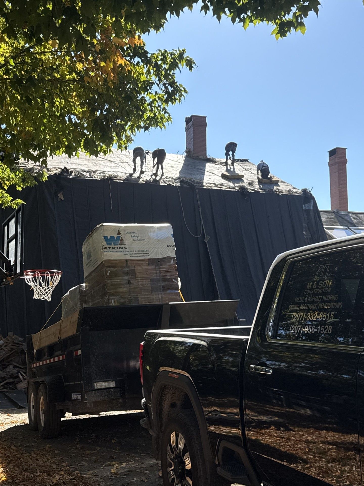 Roofing crew working on a dark roof with a truck and trailer loaded with materials.