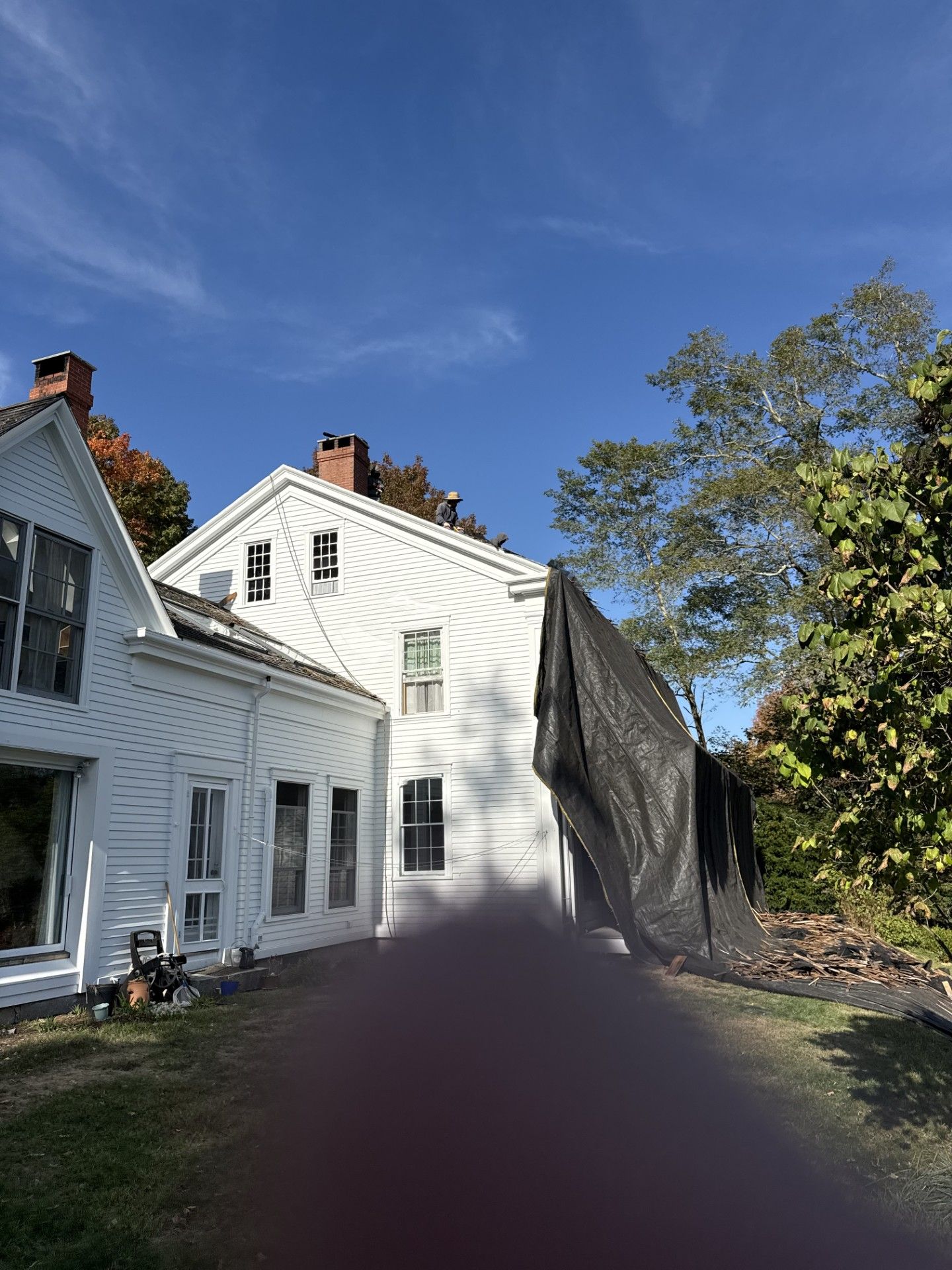 White house with a black tarp covering the side. Blue sky and autumn foliage surround it.