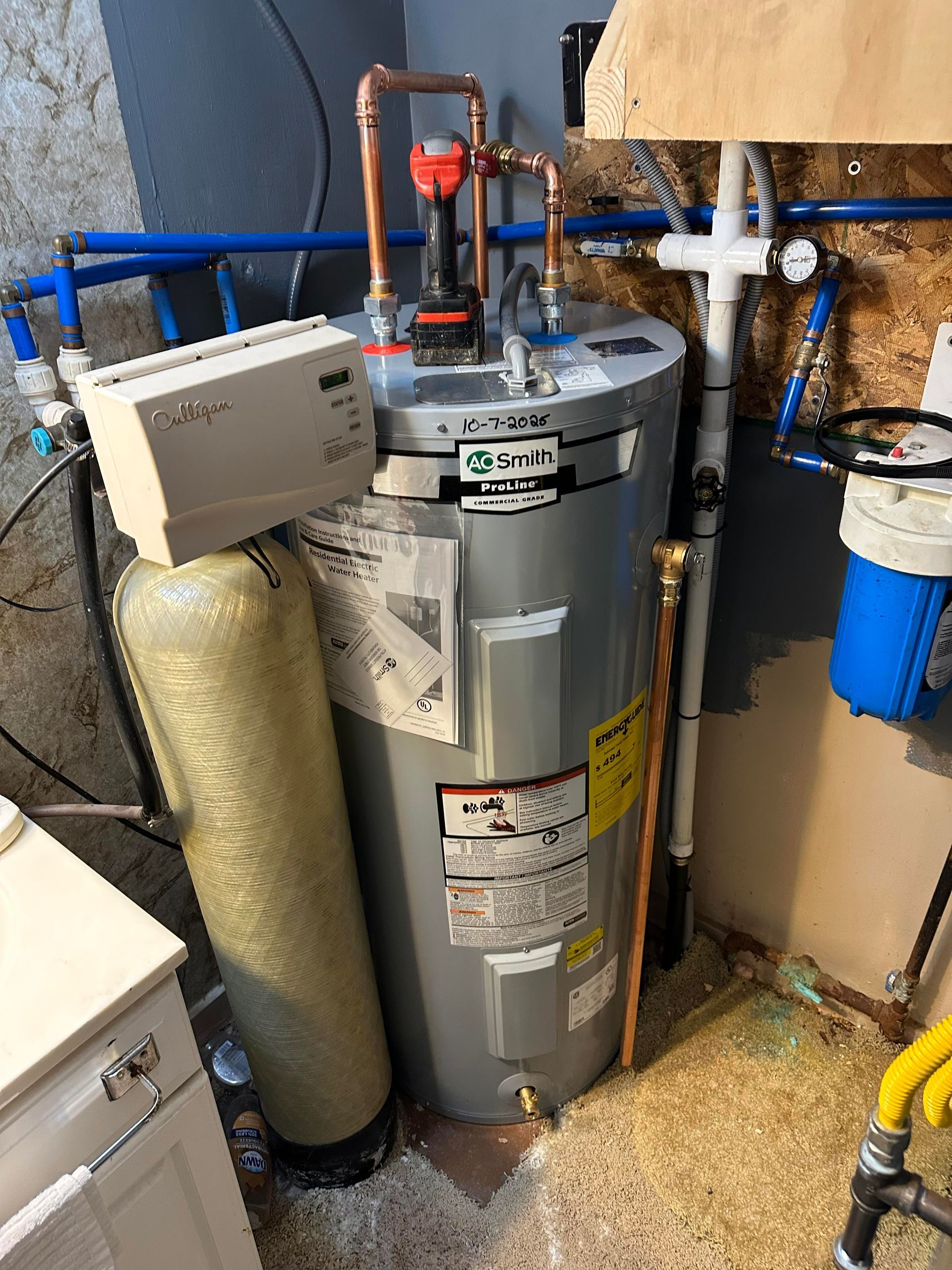 Water heater, water softener, and plumbing fixtures in a utility room. Copper and blue pipes, beige tank.