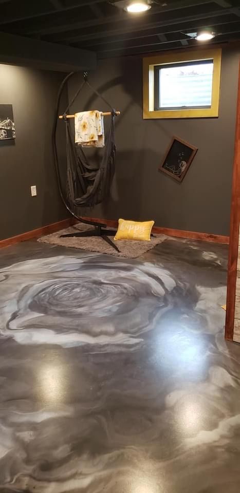 A room with dark gray walls, a hanging chair, a yellow pillow on the floor, and a floor featuring a large swirling mural.