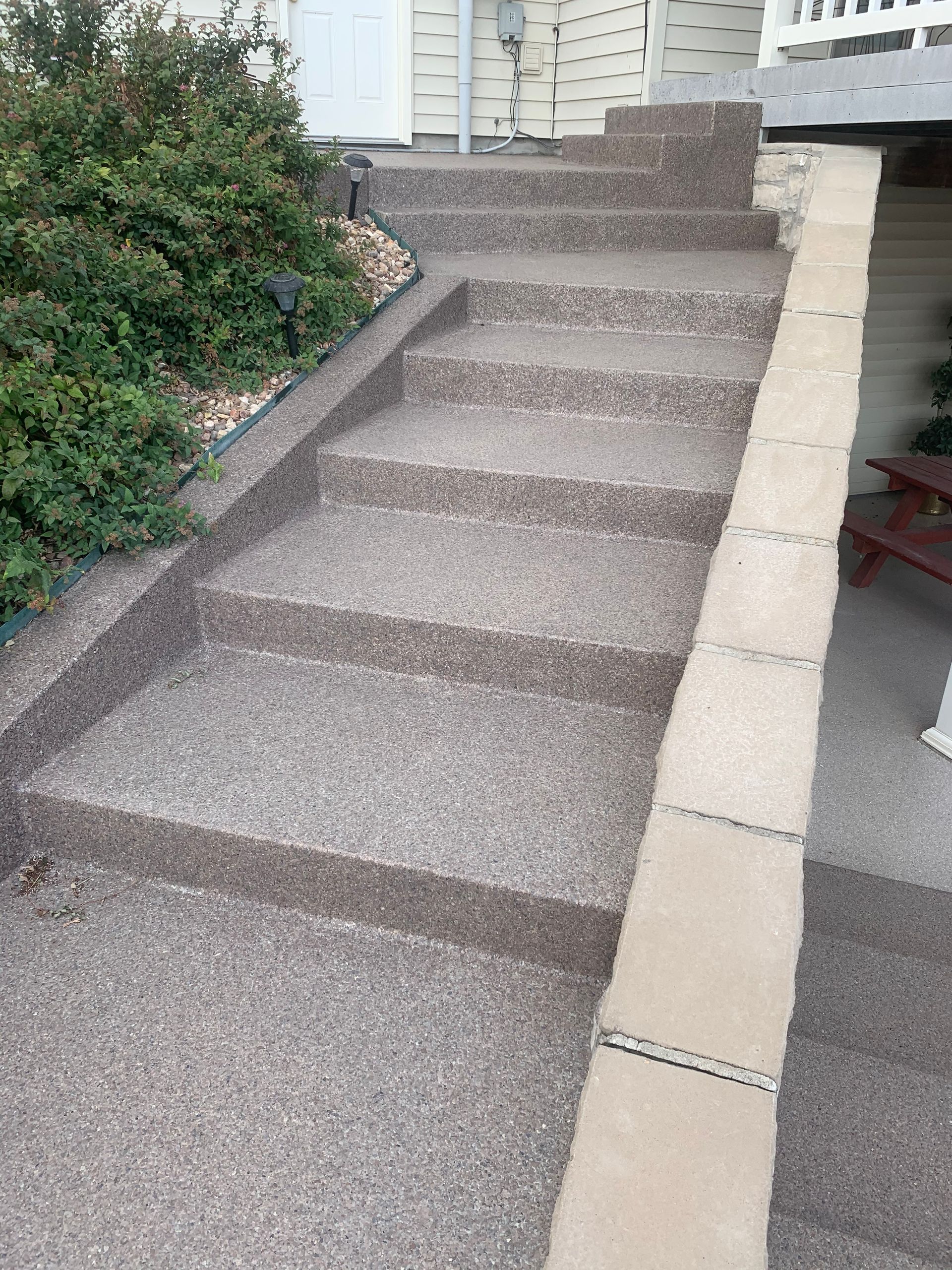 Outdoor concrete steps with a textured gray finish leading up to a house door, bordered by a beige stone retaining wall.