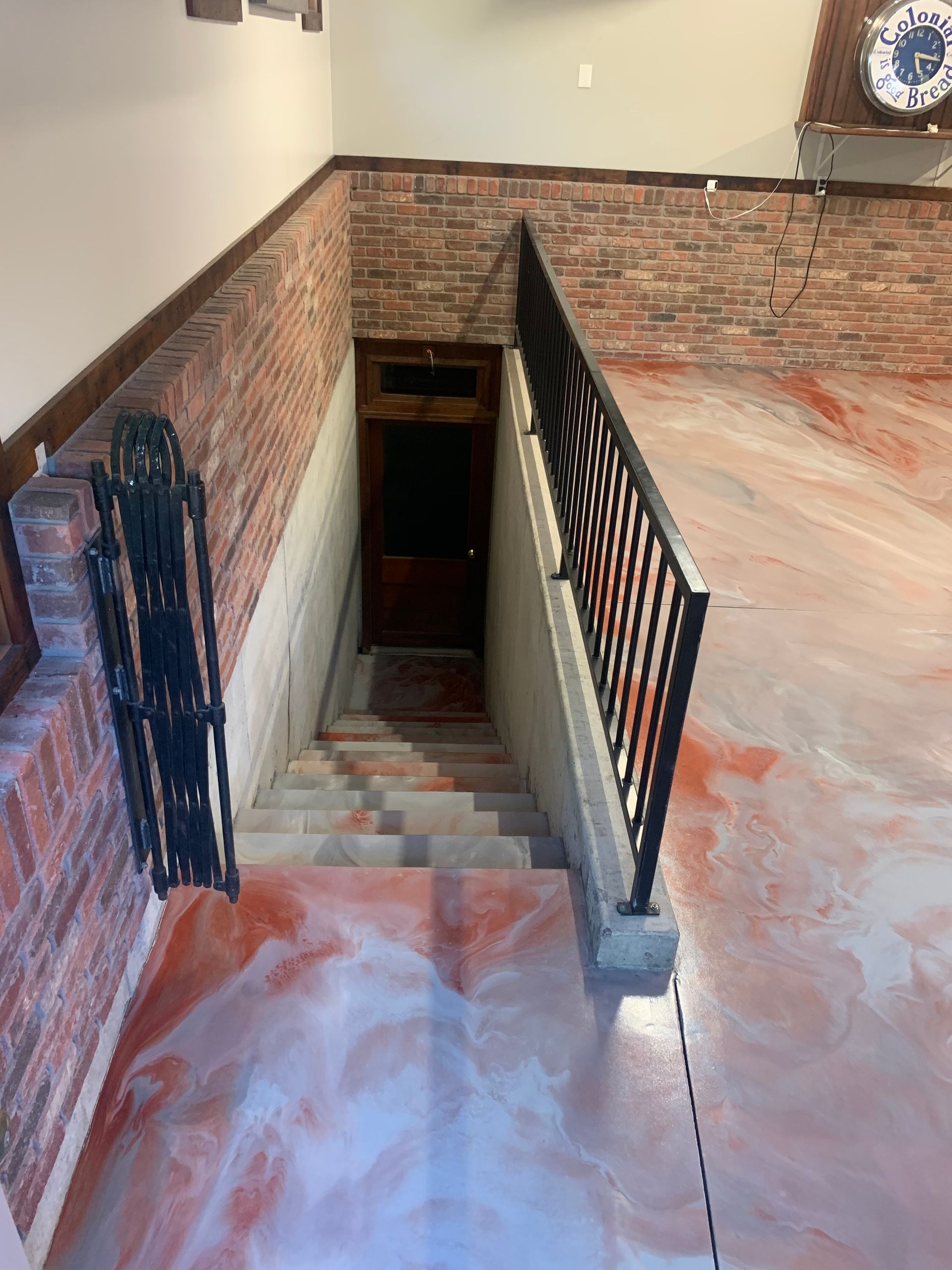 A staircase with a black iron railing leads down to a dark wooden door in a room with brick walls and swirled red flooring.