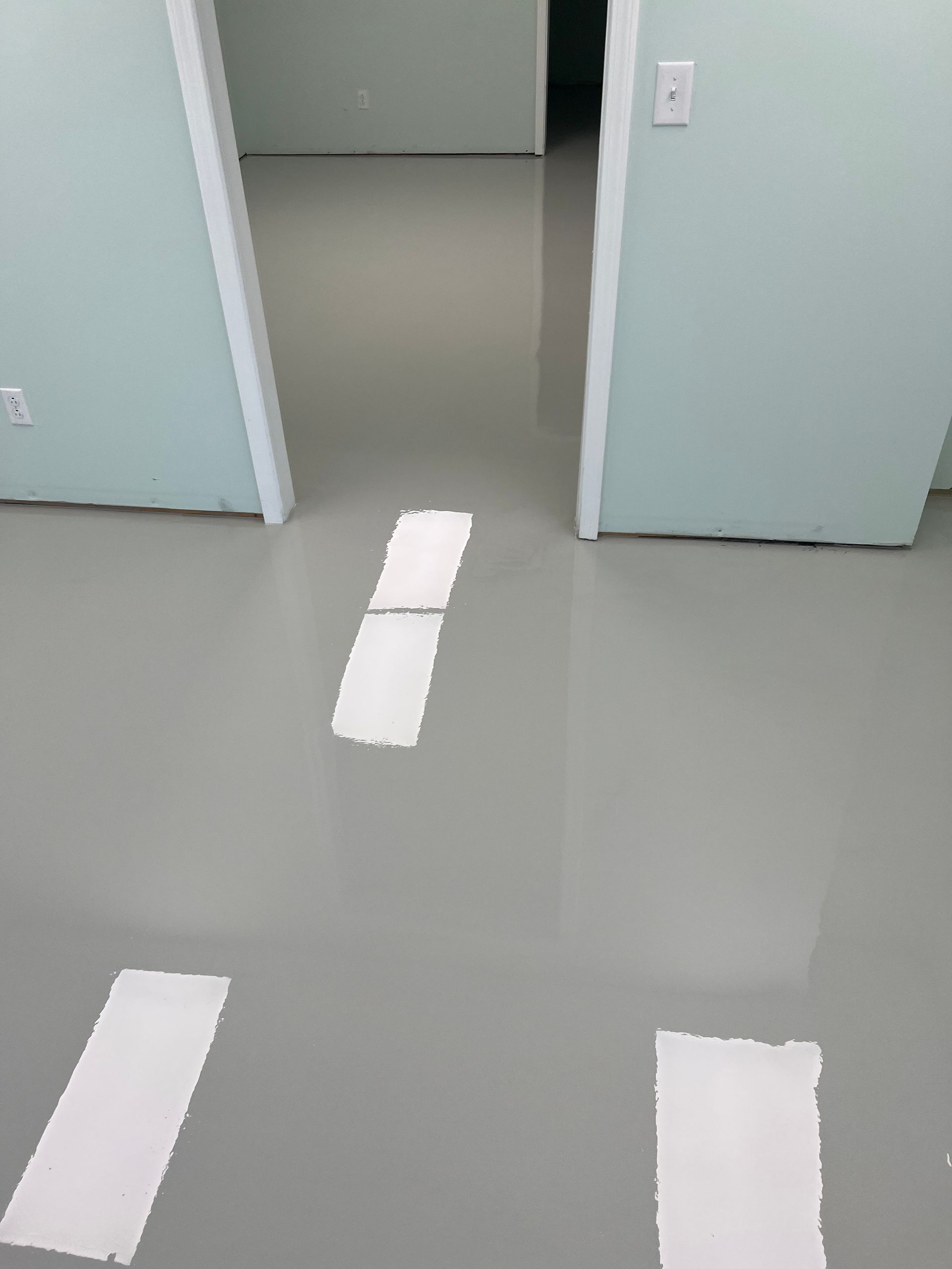 Three white rectangular floor markers painted on a smooth gray floor, leading toward a doorway in a room with light walls.