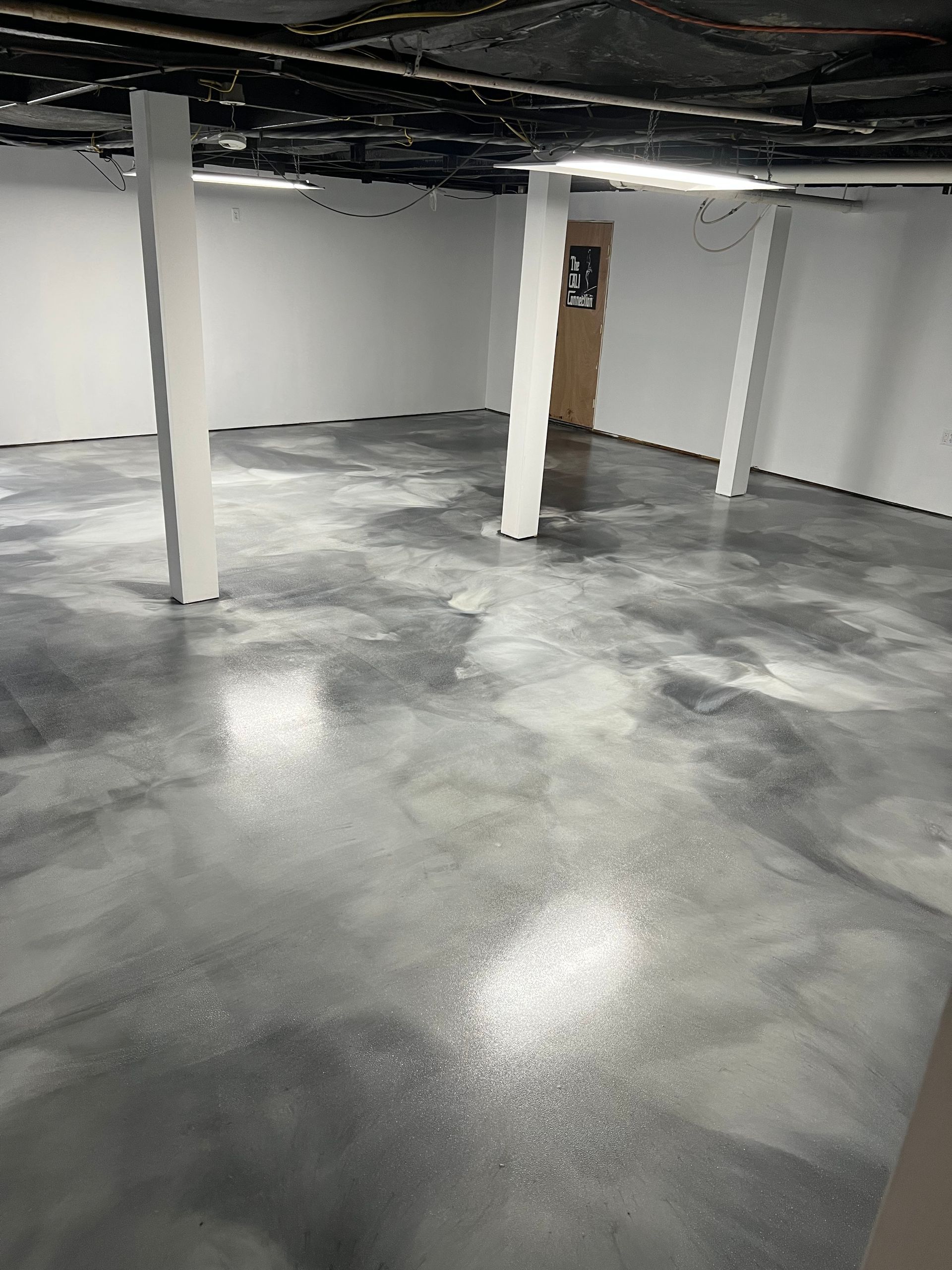 A basement room with white walls, three vertical white support posts, and a glossy, marbled grey and white epoxy floor.