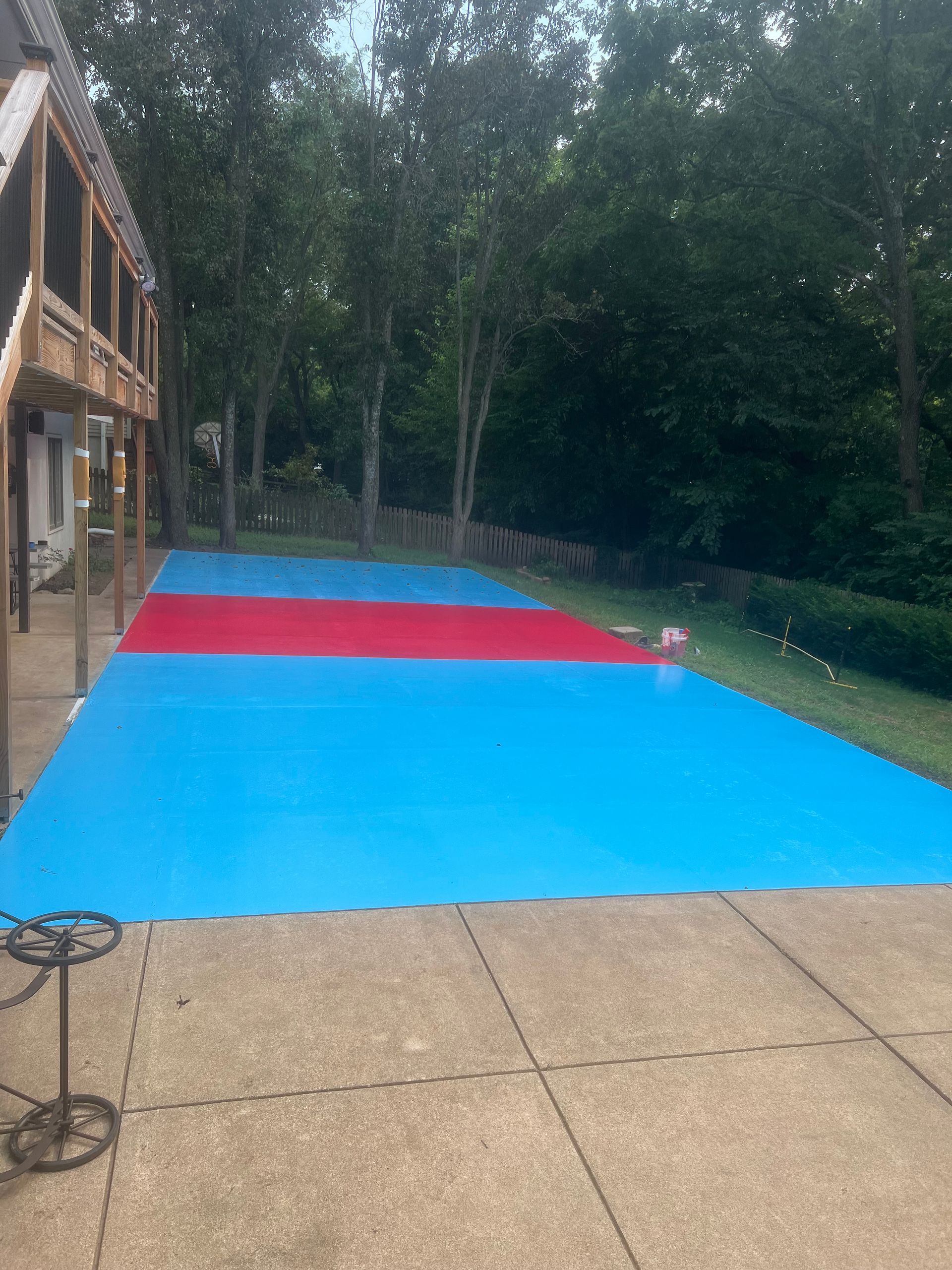 A backyard patio features a bright blue and red modular sport court surface, set near a house deck and surrounded by trees.