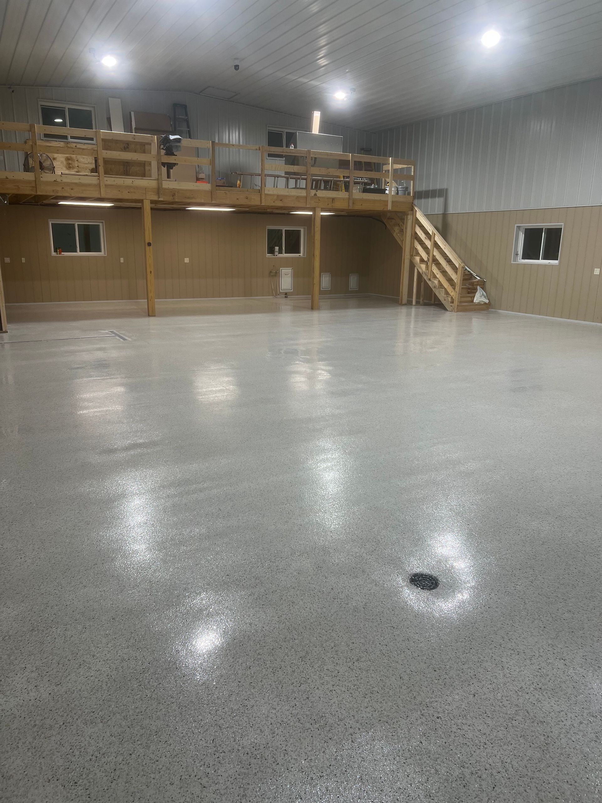 Spacious workshop with gray speckled epoxy flooring, a wooden loft with stairs, and bright overhead LED lighting.