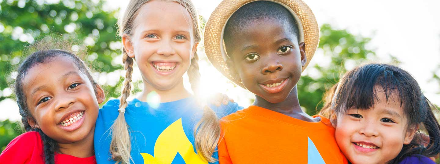 A group of young children are in a park, smiling at the camera and wearing bright clothes.